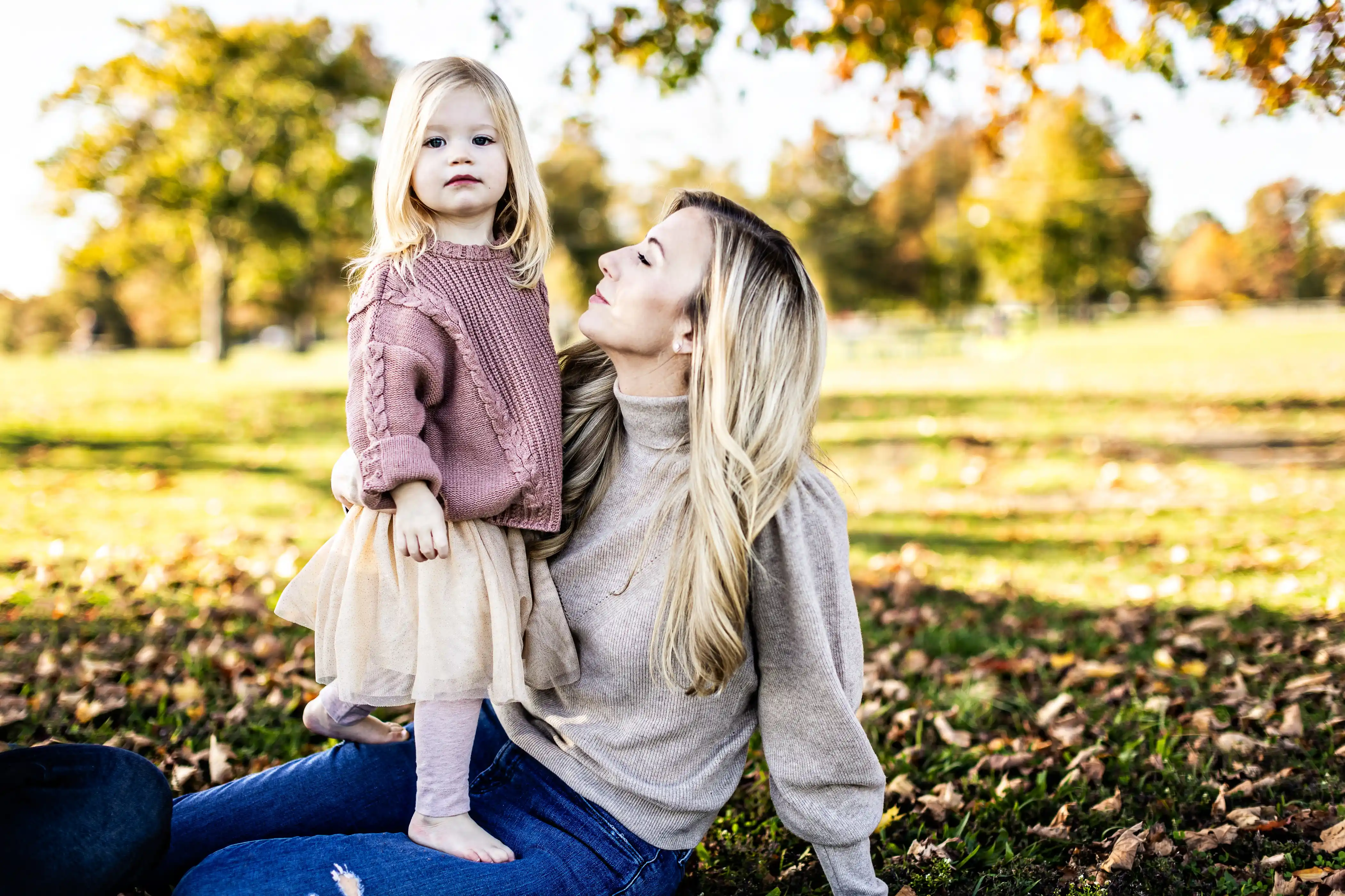A woman with long blonde hair sits on the grass in a park, looking up at a young girl standing beside her. The girl wears a pink sweater and a cream skirt. The park is filled with sunlight and autumn leaves.