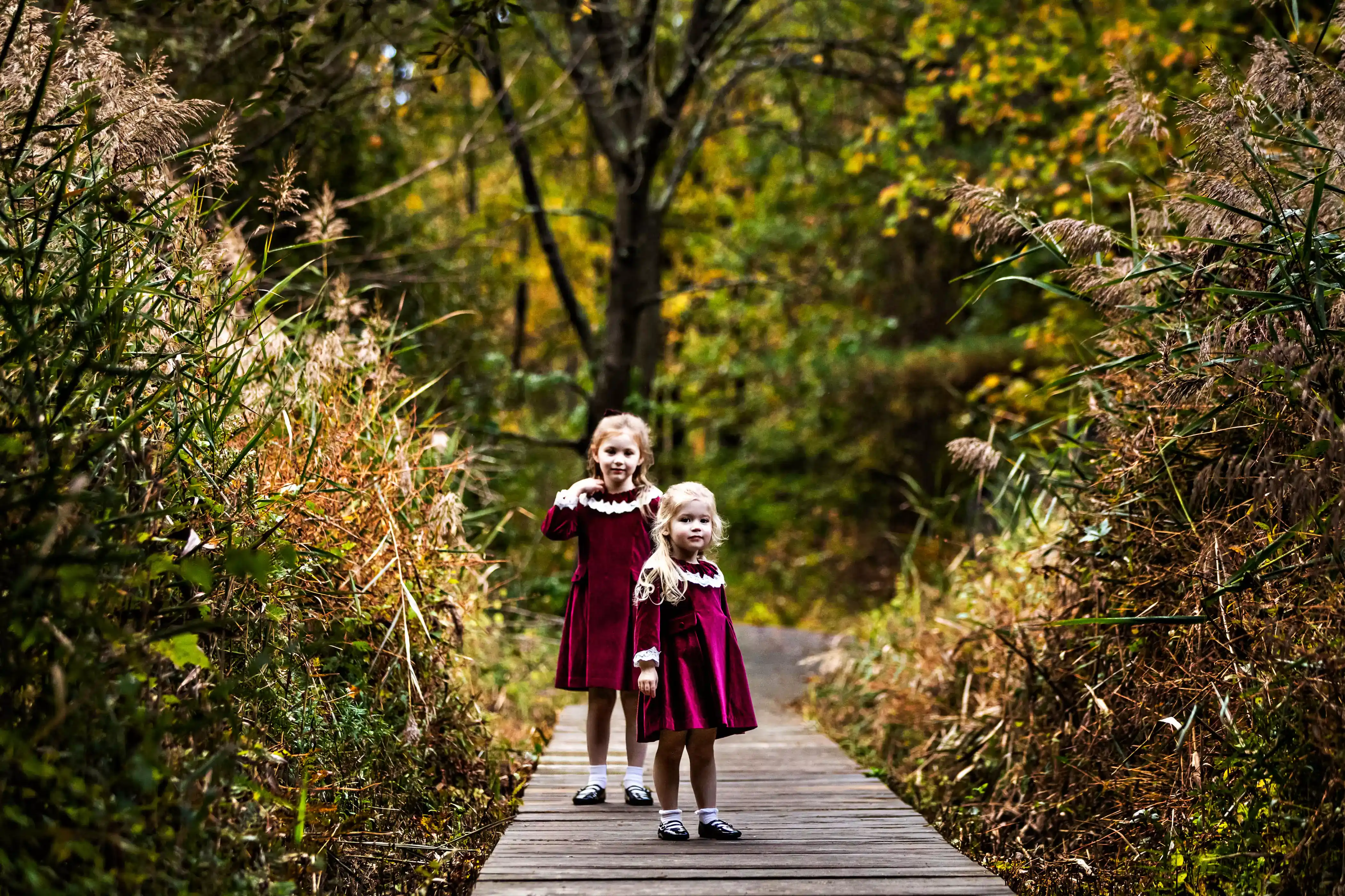 Two young girls stand on a wooden path surrounded by dense greenery. They wear matching red dresses with white collars. The taller girl waves and smiles, while the younger one looks ahead. The scene captures an autumn setting in a wooded area.
