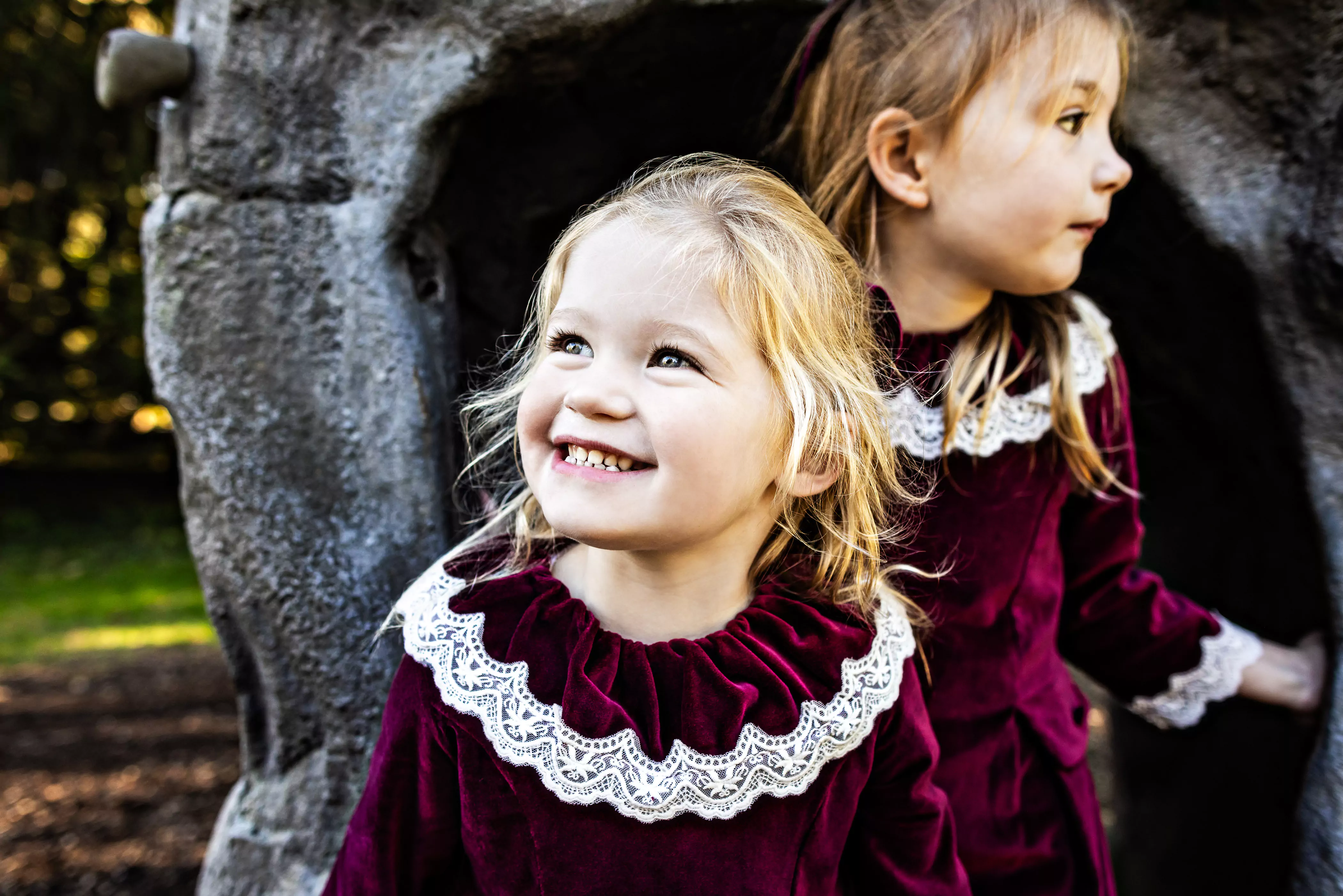 Two young girls in matching burgundy dresses with lace collars stand in front of a stone structure. One girl is smiling at the camera while the other looks to the side. The background is a mix of greenery and dappled sunlight.