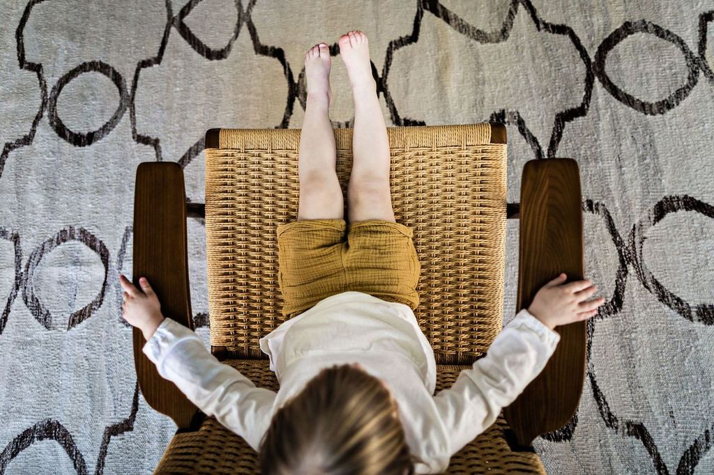 A child in a white shirt and mustard shorts lounges in a woven chair, stretching their legs out. The chair is placed on a light-colored rug with a dark geometric pattern, viewed from above.