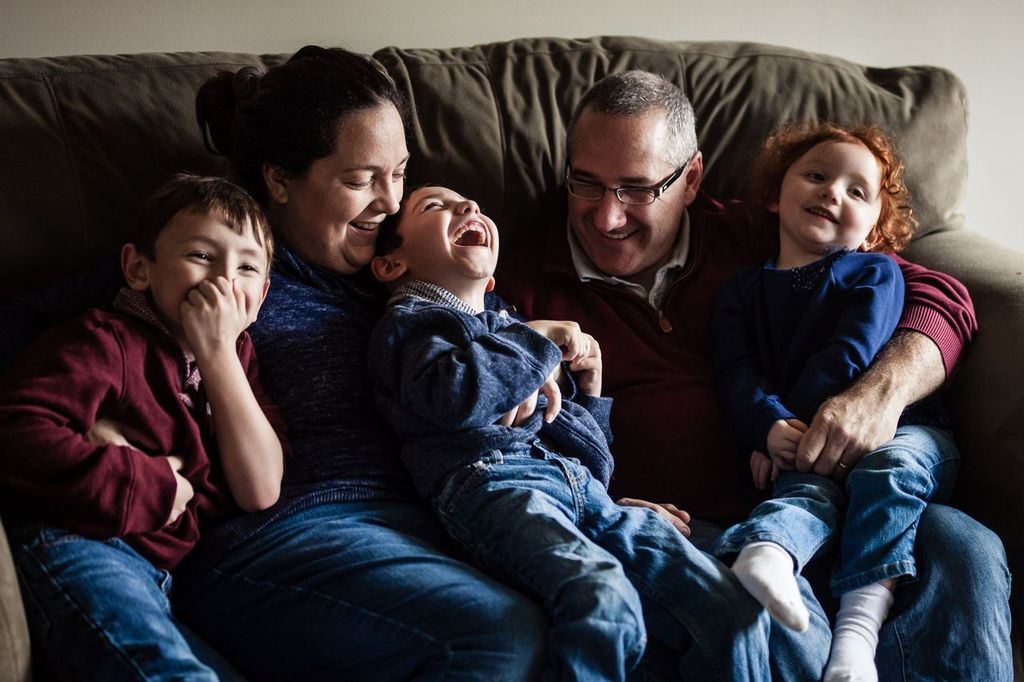 A family of five sitting on a couch, laughing and smiling. The two parents are seated in the middle, surrounded by three young children. Everyone is wearing casual clothing, and they appear to be enjoying a happy, playful moment together.