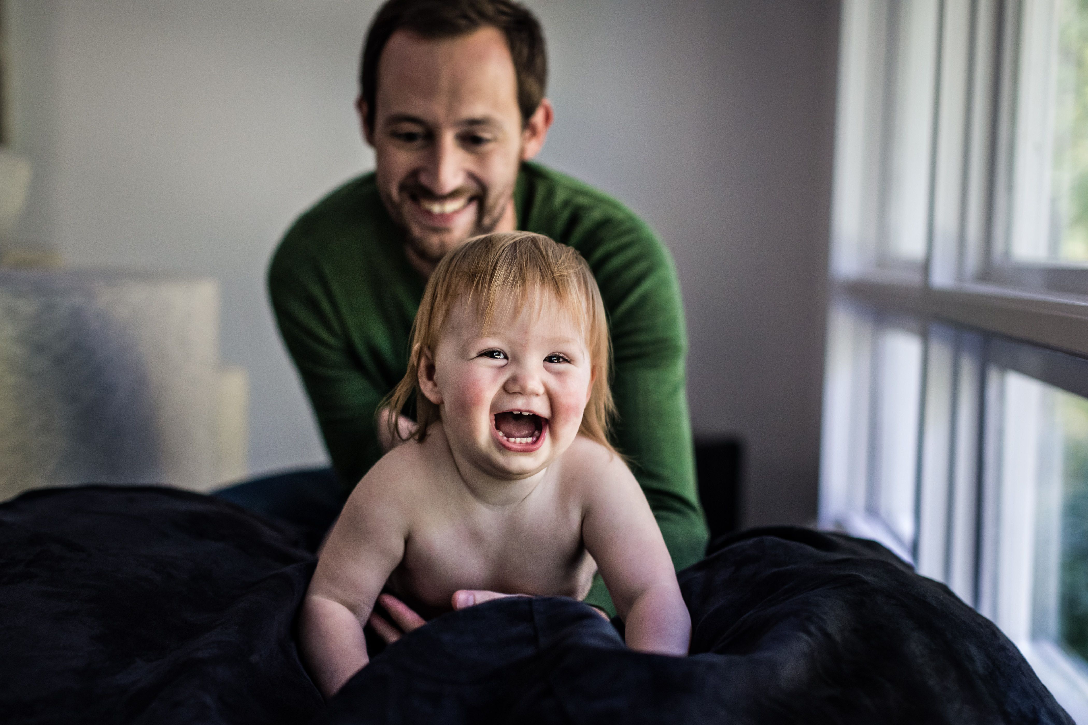 A smiling man in a green sweater playfully holds a happy baby with light hair who is lying on a dark blanket. The setting is bright, with large windows in the background, creating a warm and joyful atmosphere.