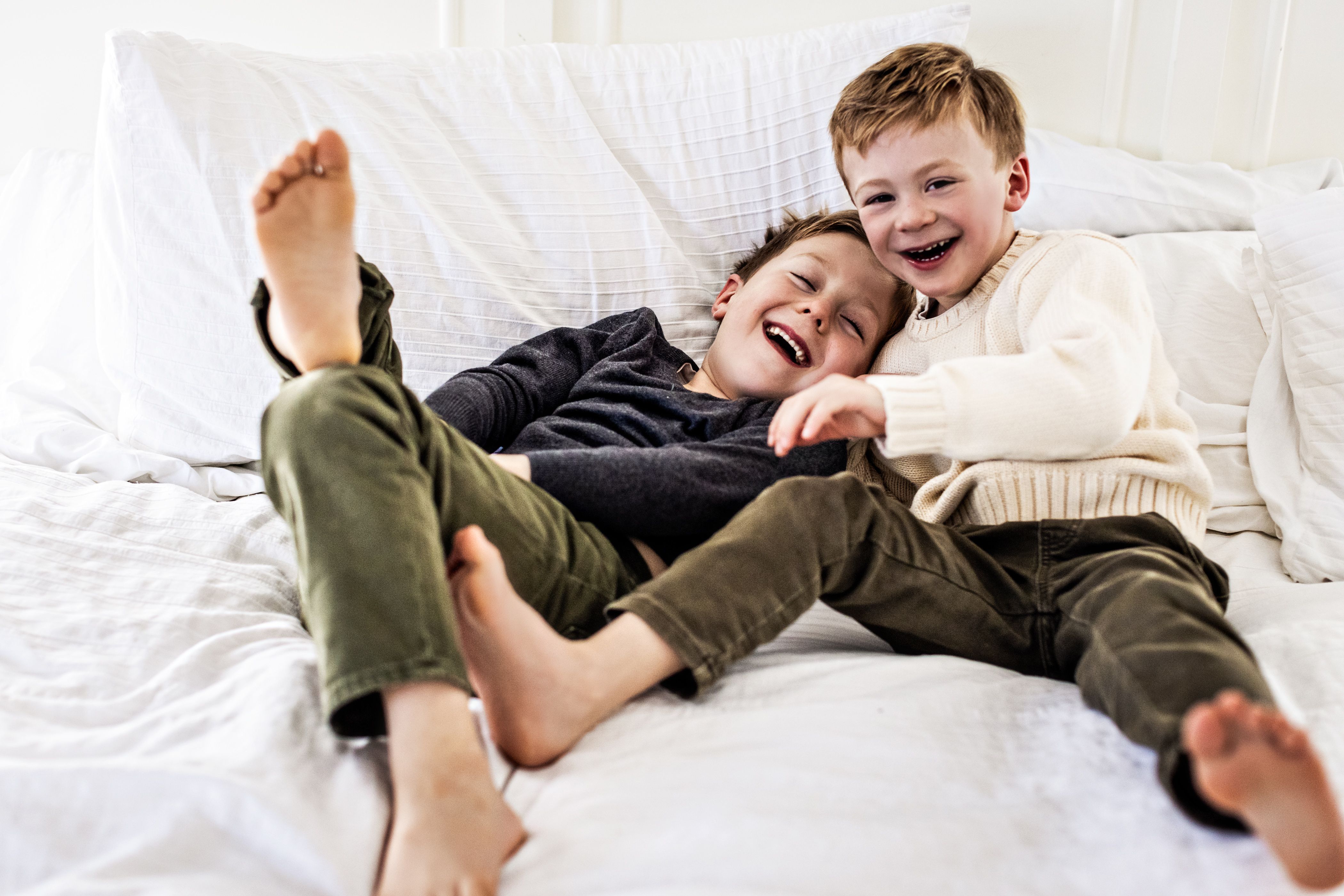 Two young boys laughing together on a bed, dressed in casual sweaters and pants. One has light brown hair and the other light blonde. They lie on white bedding, displaying a joyful and carefree moment.