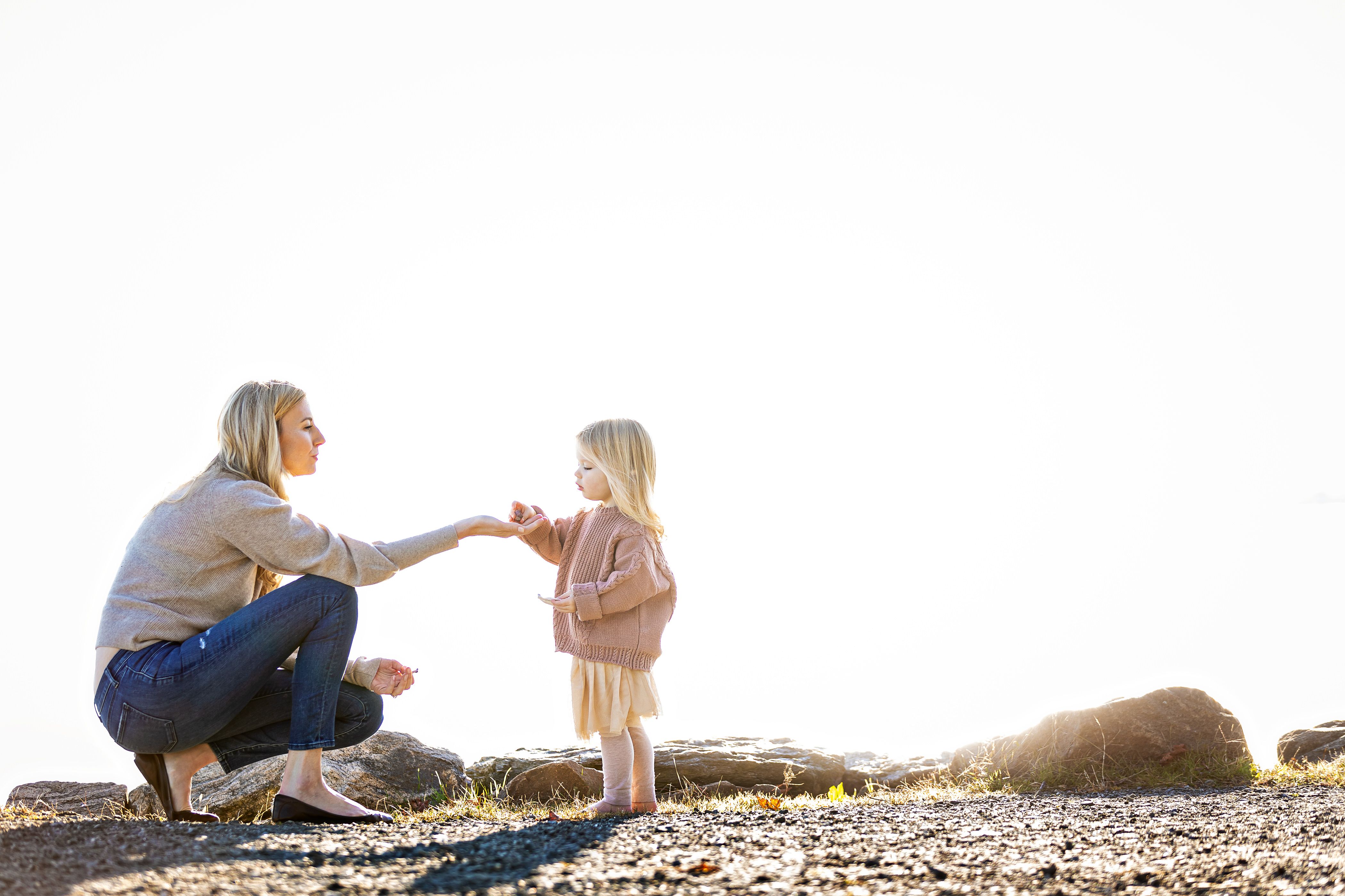 A woman crouches to hold hands with a child by a rocky shoreline, both wearing casual clothes. The bright, sunny background creates a soft, serene atmosphere.