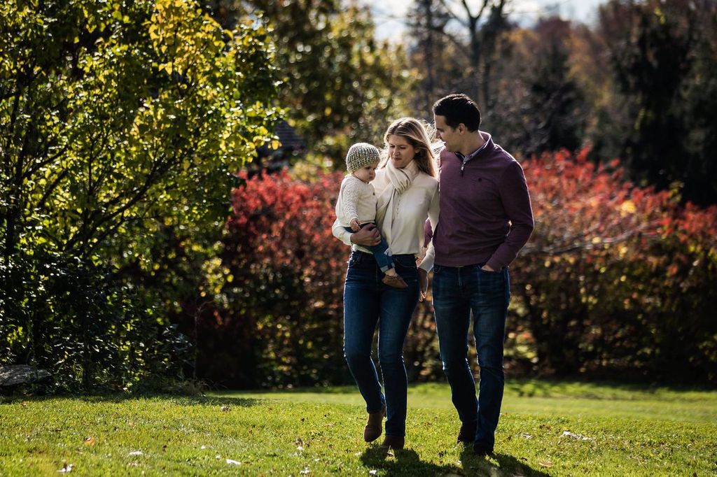 A couple is walking in a park holding a child. They walk on a grassy area with vibrant autumn foliage, including red and green trees, in the background. The child is wearing a knit hat. The scene conveys a warm, family outing in nature.