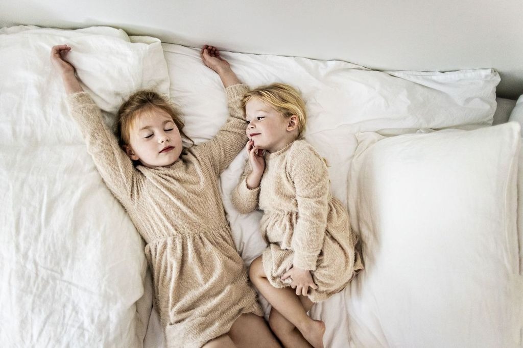 Two young children wearing matching beige outfits lie on a bed with white sheets and pillows. One child is sleeping with arms stretched above their head, while the other, awake, looks at the sleeper with a gentle expression.