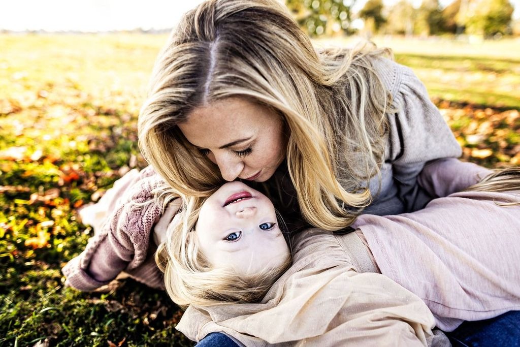 A woman with long blonde hair kisses a smiling child on the forehead as they lie on grass sprinkled with autumn leaves. The scene evokes warmth and tenderness in an outdoor setting.