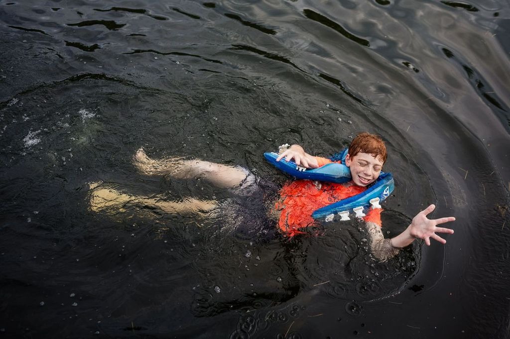 A person wearing a bright orange life vest is floating in dark water, looking upward with one arm extended out. The persons eyes are closed, and they appear to be treading water. Ripples surround them on the waters surface.