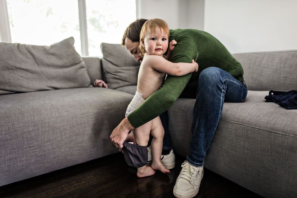 A person wearing a green sweater helps a toddler in a diaper put on pants. They are by a gray couch in a well-lit room. The child looks towards the camera.