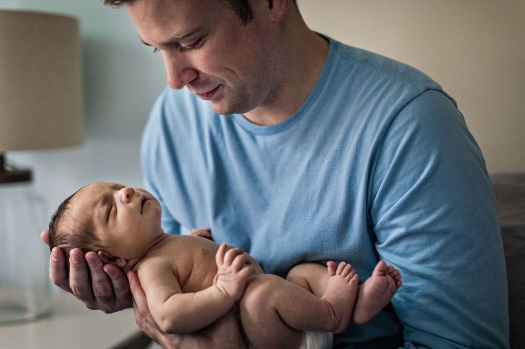 A man in a blue shirt gently cradles a sleeping baby in his arms. The setting is calm and homely, with a lamp in the background. The man gazes tenderly at the baby.