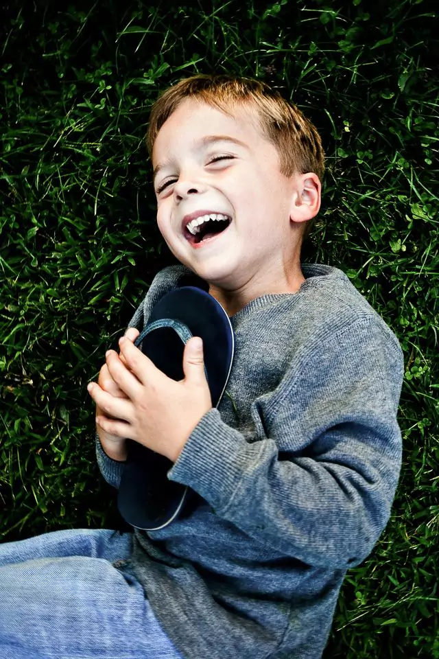 A young boy lies on green grass, laughing joyfully. He holds a black shoe to his chest and wears a gray sweater and blue pants. His eyes are closed, and his hair is light brown.