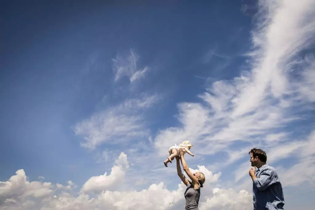 A woman joyfully lifts a baby into the air under a bright blue sky with scattered clouds. A man stands nearby, smiling and watching them.