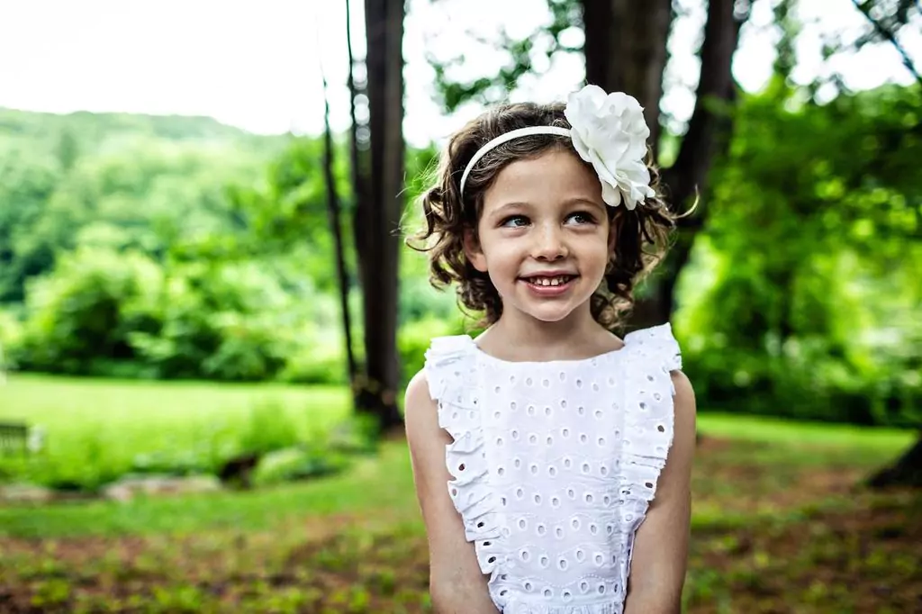 A young girl with curly hair, wearing a white dress with eyelet details and a large white flower headband, smiles while standing in a garden. The background is lush and green, with trees and grass visible.