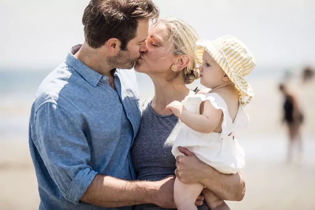 A couple is kissing on a beach while the woman holds a baby. The man is wearing a blue shirt, and the woman is in a gray top. The baby, in a white outfit and striped hat, looks away. The background shows the ocean and blurred figures.