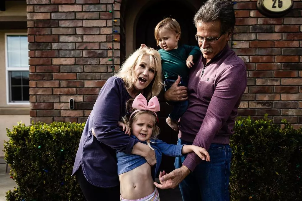 A woman and a man stand in front of a brick house. The woman is playfully holding a young girl with a pink bow, who is pulling up her shirt. The man holds a toddler. They all appear happy and lively.