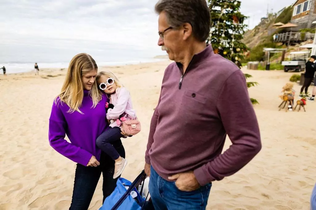 A woman in a purple sweater holds a young child wearing sunglasses and pink earmuffs on a sandy beach. A man in a maroon sweater stands nearby. Beach houses and people are visible in the background under a cloudy sky.