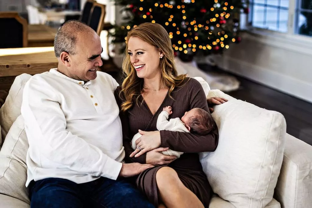 A man and woman sit happily on a couch, smiling at each other. The woman cradles a sleeping baby. A decorated Christmas tree with lights is in the background, creating a festive atmosphere.