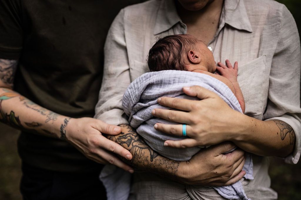 A close-up image of two adults holding a newborn wrapped in a soft blanket. One adult is wearing a light shirt and a blue ring, while the other is in a dark shirt, both showing tattooed arms gently supporting the baby.