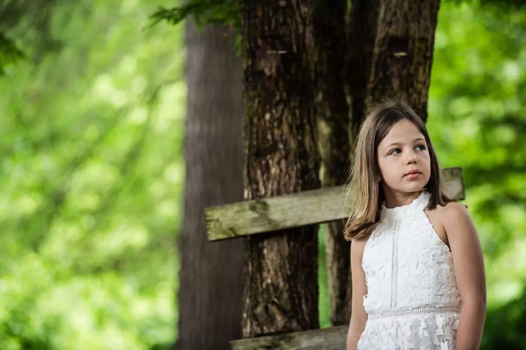 A young girl in a white dress stands in a forested area, looking to her left. She is next to a wooden fence with lush green trees in the background.