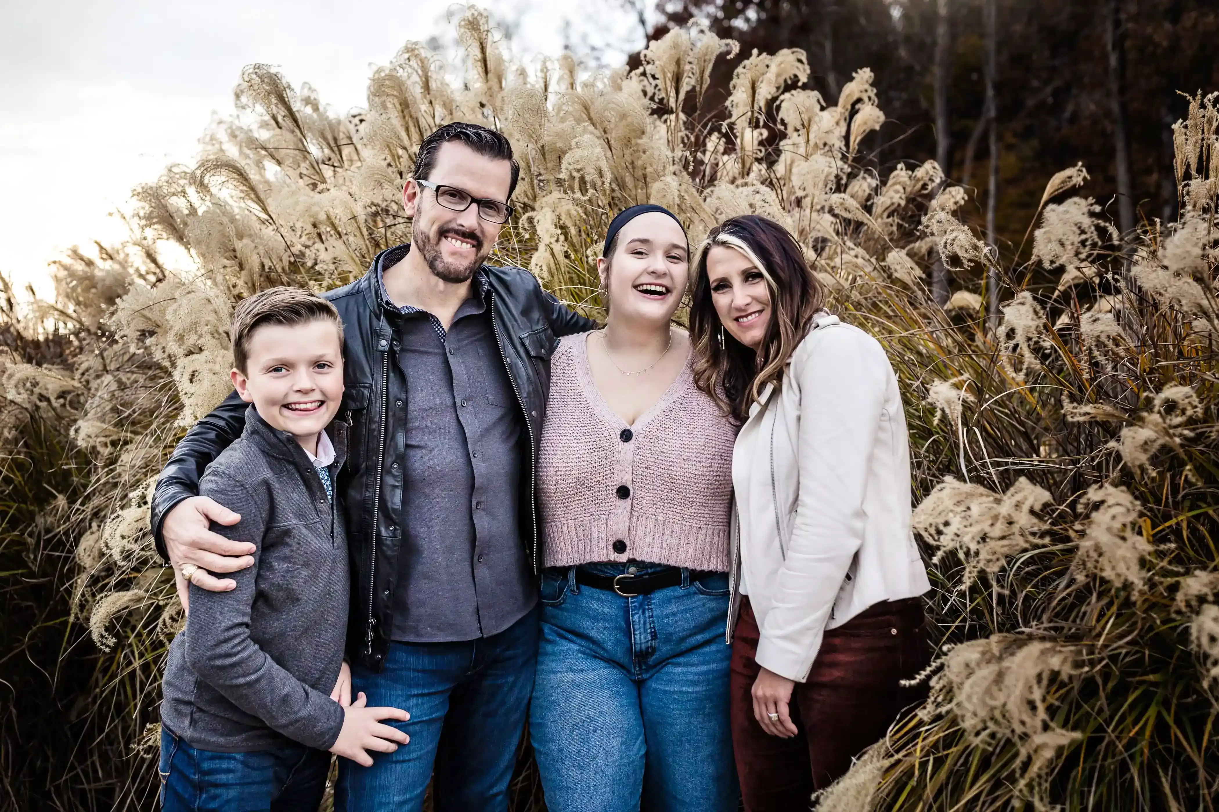 A family of four stands together smiling in front of tall, feathery grass. The father is wearing a leather jacket, the mother is in a white jacket, the daughter is in a pink cardigan, and the son is in a gray sweater.