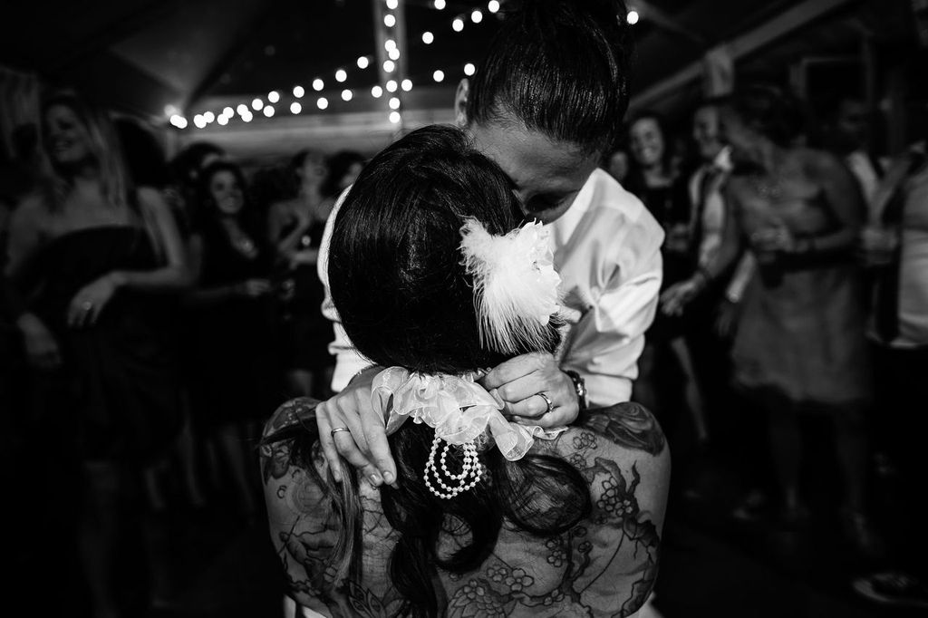 Artistic Composition02 Two women embrace closely on a dance floor, surrounded by people. One wears a lace dress with a feather hair accessory and pearls; the other wears a white shirt. String lights hang overhead, creating a festive and intimate atmosphere.