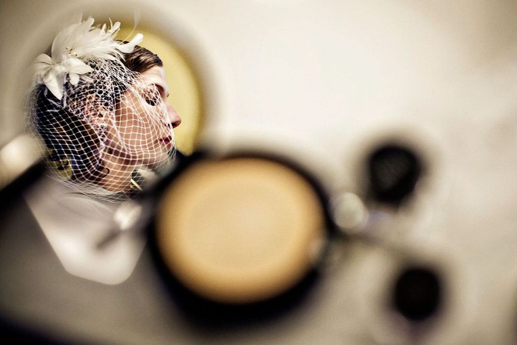 Blurred Imagery A woman wearing a white netted fascinator with feathers is reflected in a mirror, her eyes closed and face calm, surrounded by blurred makeup items in the foreground.