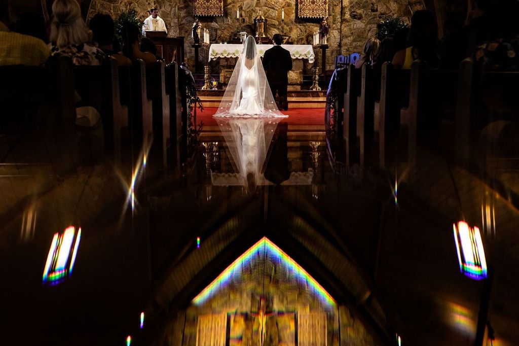 Dramatic Lights A bride and groom kneel at the altar in a dimly lit church, with their reflections visible on the polished floor and colorful light from stained glass adding a vibrant touch. Guests are seated in pews on either side.