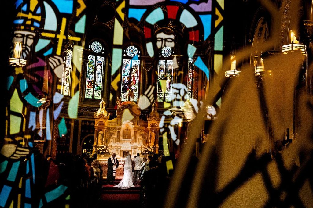 Capturing Details A wedding ceremony takes place in a church, seen through colorful stained glass windows that show religious figures, creating a layered, vibrant effect over the bride, groom, and guests at the altar.