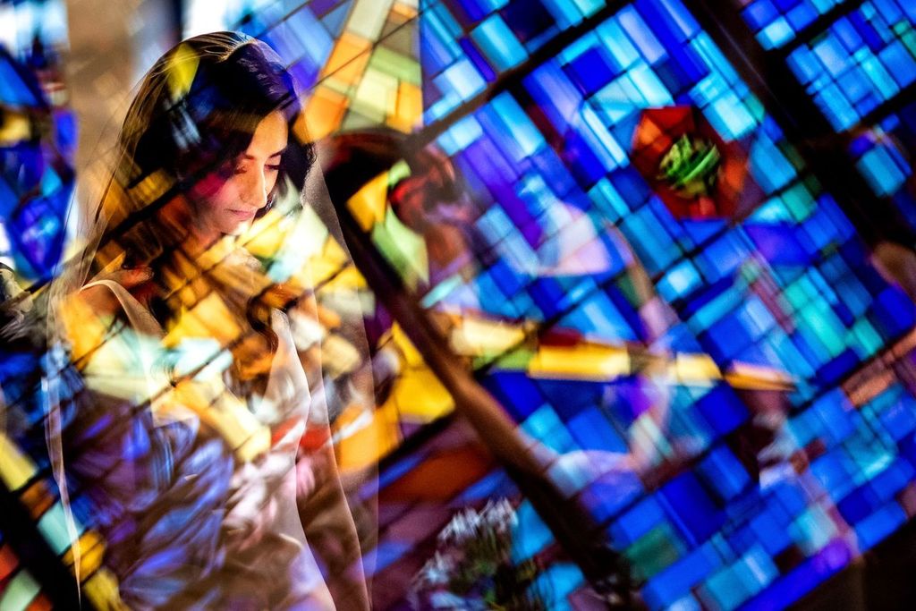Dramatic Lights A woman stands with her eyes closed, bathed in colorful light streaming through a vibrant stained glass window, creating a layered, reflective effect around her.