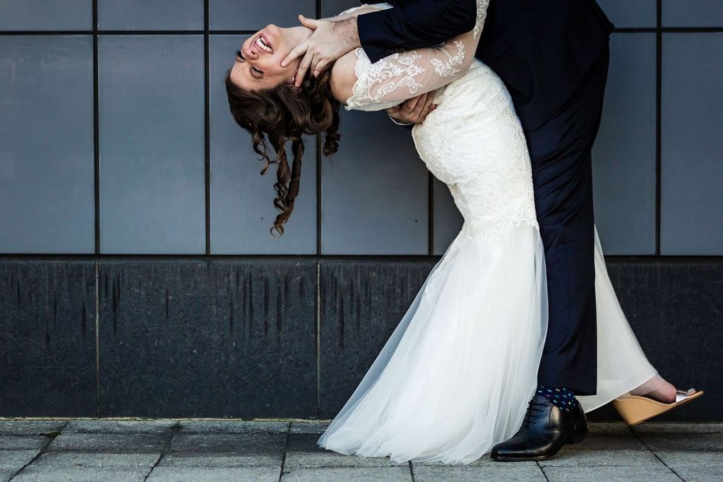 Creative Posing 02 A bride in a white lace dress laughs as she is playfully dipped backward by a groom in a dark suit, standing on a sidewalk in front of a tiled wall.