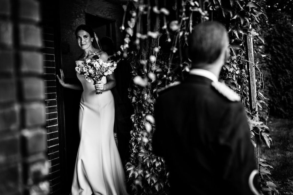 Creative Posing 03 A bride in a white dress and holding a bouquet steps out of a doorway, smiling, while a man in formal attire stands in the foreground, his back to the camera. The scene is surrounded by foliage.