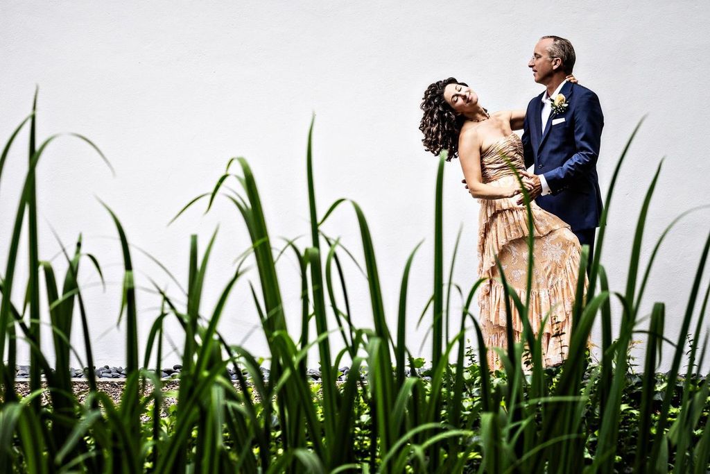 Creative Posing A couple dressed formally pose together in front of a plain white wall, with tall green grass in the foreground. The woman leans back gracefully, supported by her partner, who looks at her and smiles.