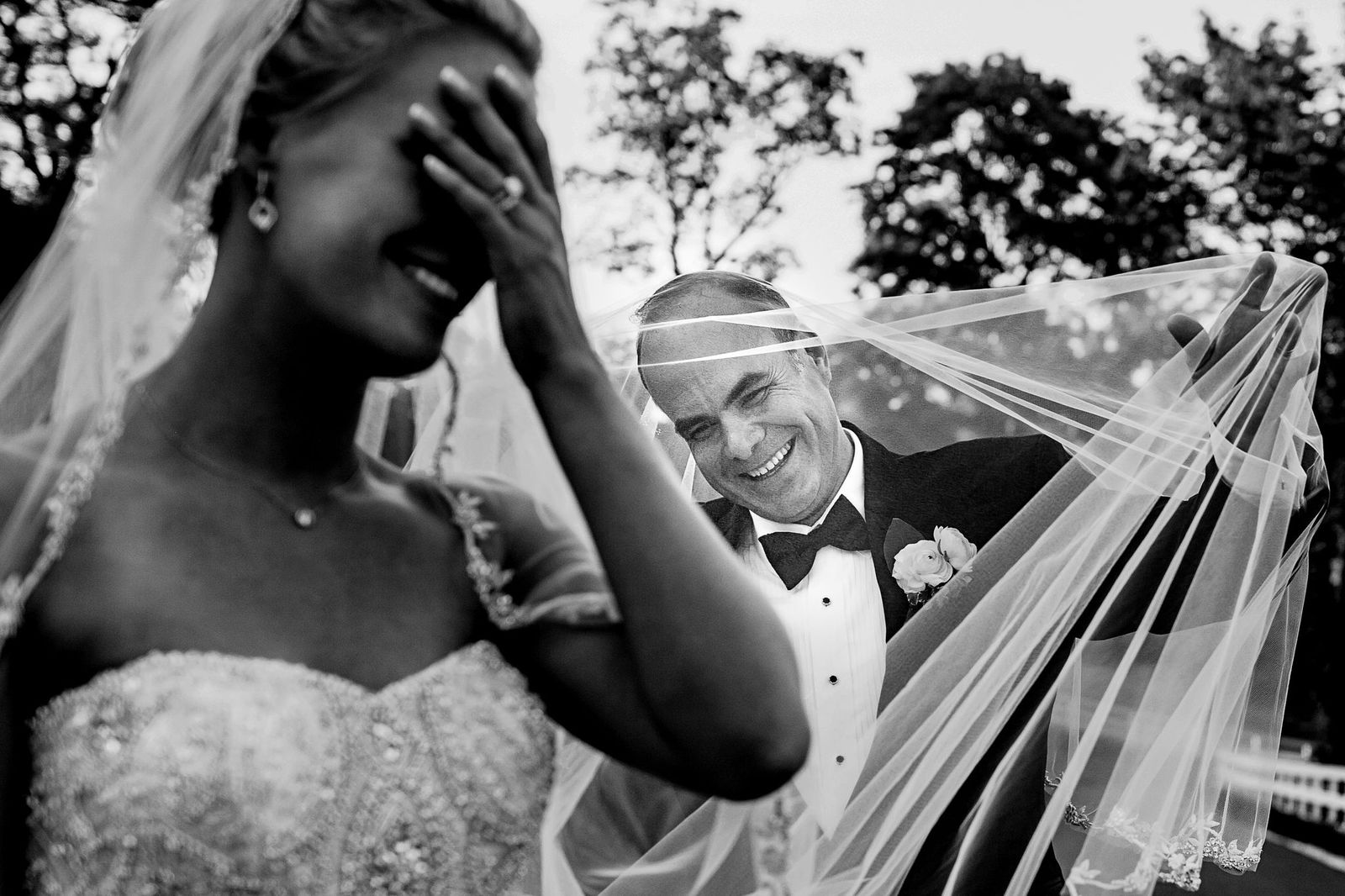 Editorial Wedding 03 A bride laughs with her hand over her face as a smiling man in a tuxedo, possibly her groom, playfully holds her wedding veil. The scene is joyful and candid, set outdoors with trees in the background.