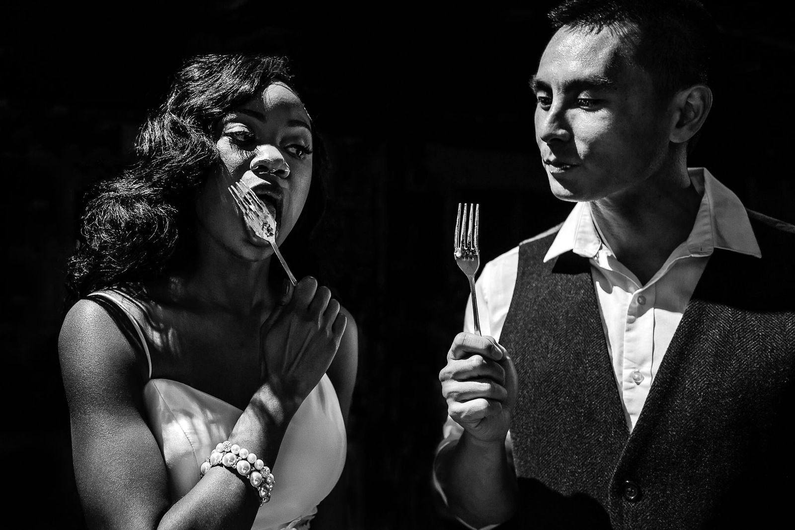 Editorial Wedding A woman in a dress eats food from a fork while a man in a vest and collared shirt looks at her, both standing in dramatic lighting against a dark background.