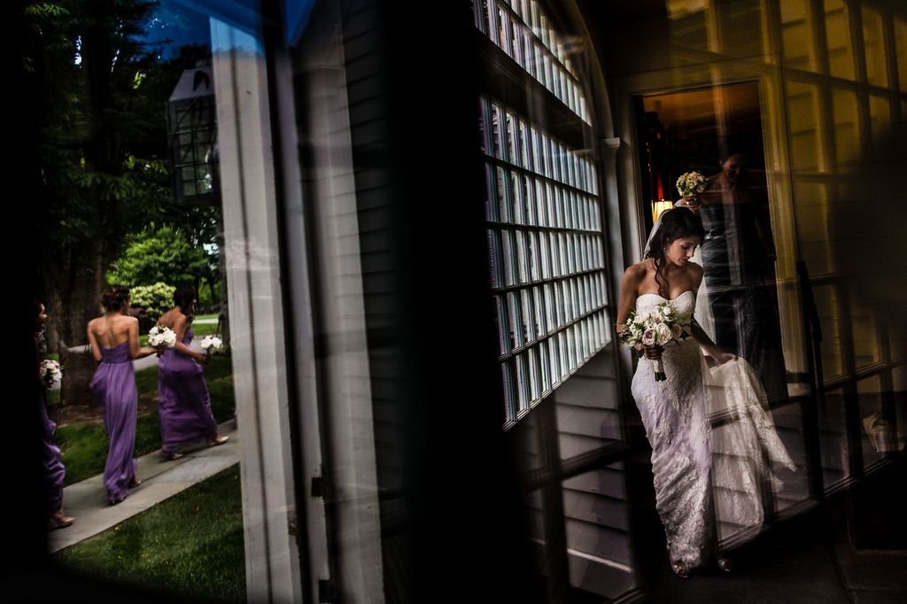 Fashion Influence A bride in a white gown stands indoors near large windows, holding her dress and bouquet. Reflected in the glass, bridesmaids in lavender dresses walk outside on a garden path.