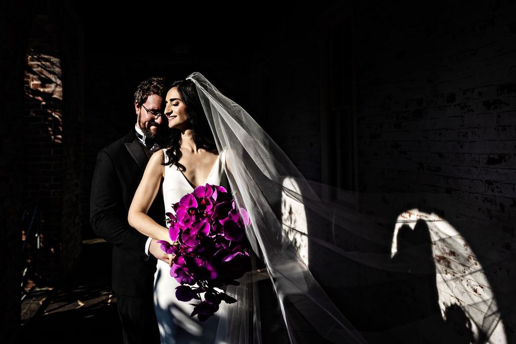 Good Lighting and Editing A bride in a white dress and veil holds vibrant purple orchids while standing with her groom in a dark, shadowy setting. Sunlight highlights the couple, creating dramatic light and shadow effects on the wall behind them.