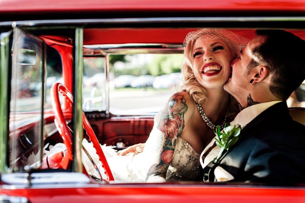 Purpose of Editorial Wedding Photography A joyful bride with tattoos and a netted veil laughs while sitting in a red vintage car next to her smiling groom, who is kissing her cheek. Sunlight spills into the car, highlighting their happiness.