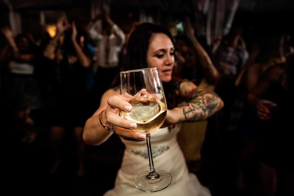 Purpose of Editorial Wedding Photography01 A woman in a white dress holds a glass of white wine toward the camera at a lively event, with people dancing and celebrating in the blurred background.