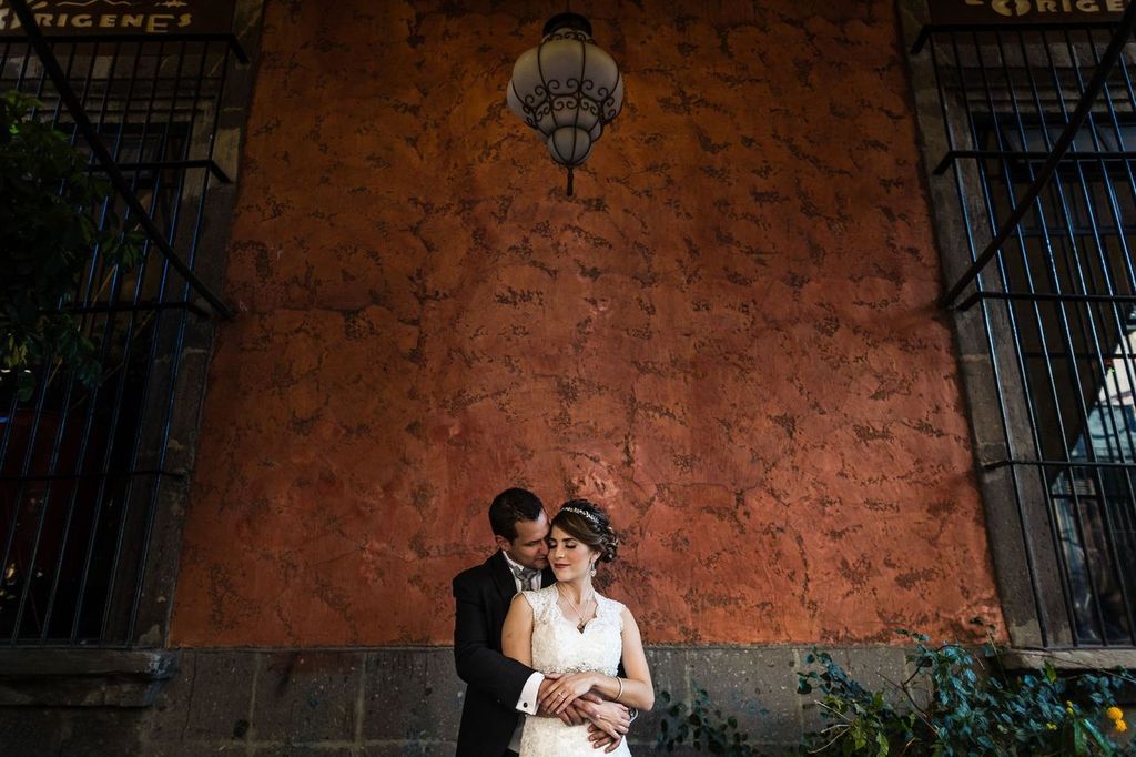 Storytelling A bride and groom embrace lovingly in front of a textured, orange-red wall with a vintage lamp above them. The bride wears a lace dress and headpiece; the groom, in a suit, holds her from behind. Greenery frames the bottom corners.