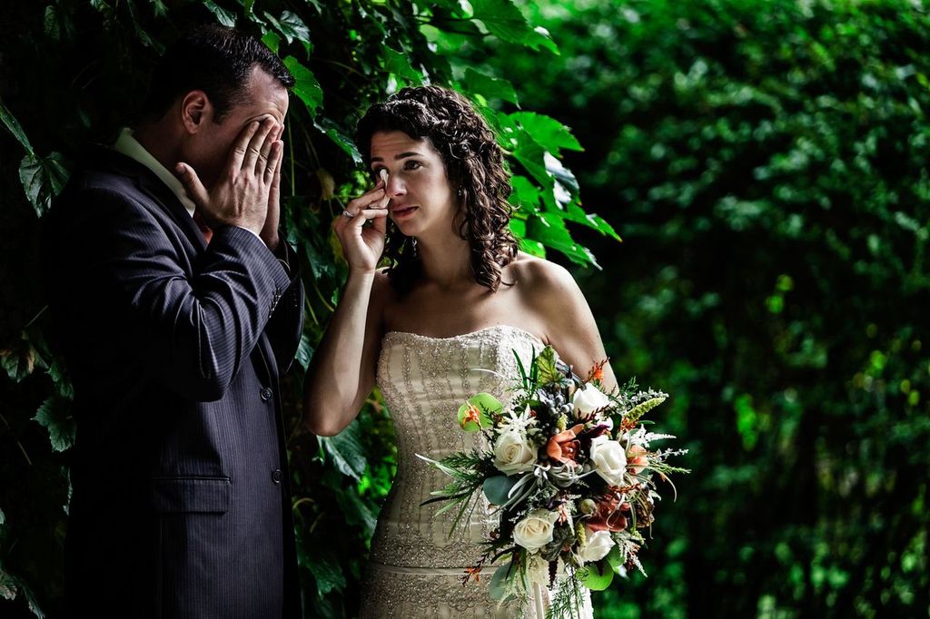 jagstudios-stephanie-dex-wedding-look-park-florence-ma-photography-032 A bride holding a bouquet and a groom in a suit stand together outdoors, both wiping tears from their eyes, surrounded by lush green foliage.