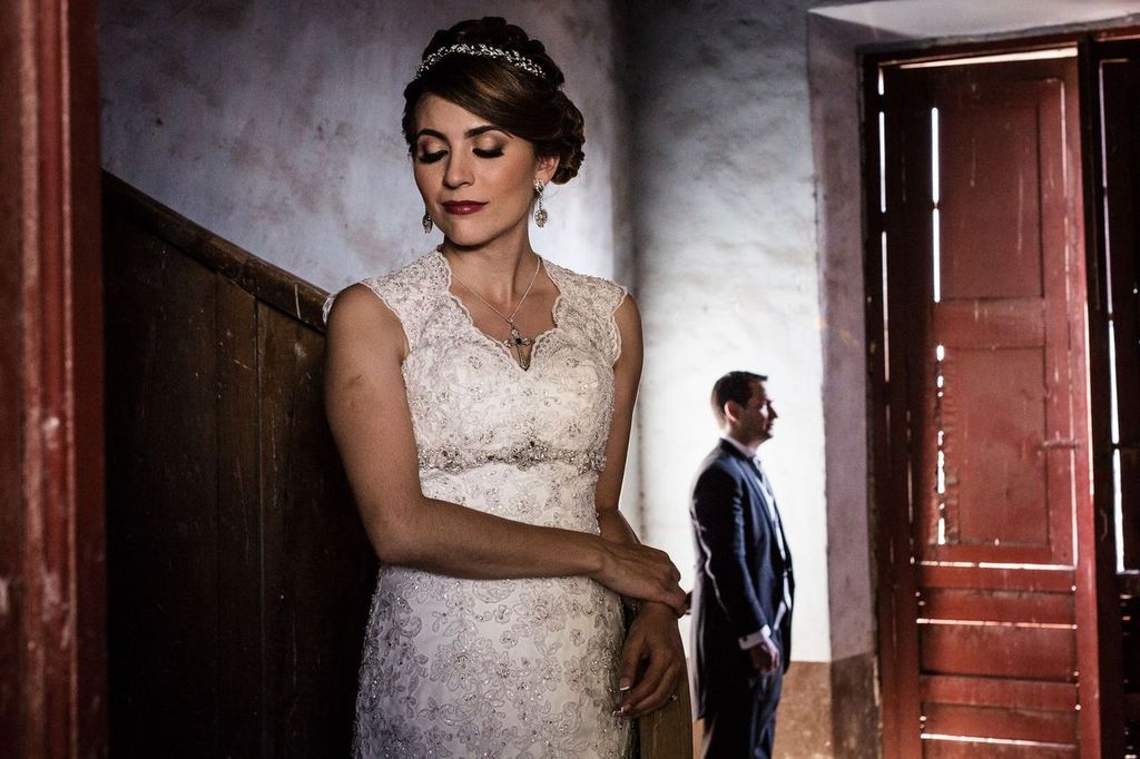 storytelling02 A bride in a white lace dress stands with eyes closed by a wooden wall, while a groom in a suit stands in the background near open double doors letting in sunlight.